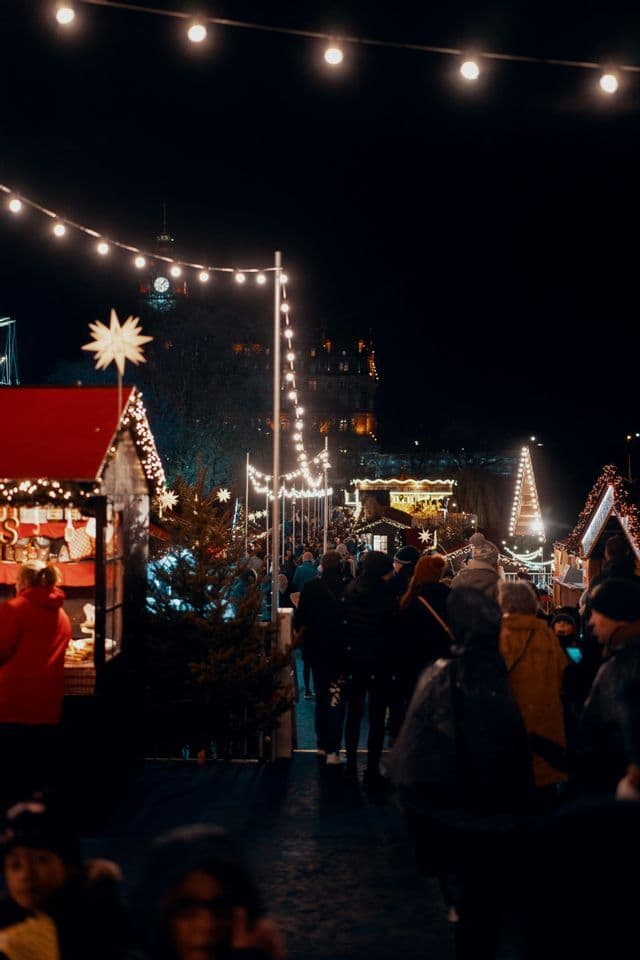 Una folla di persone passeggia in un mercatino di Natale all'aperto di notte, con bancarelle e vialetti illuminati da luci natalizie.