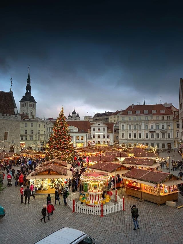 Un vivace mercatino di Natale anima una piazza lastricata con un grande albero decorato, bancarelle e una giostra al crepuscolo.