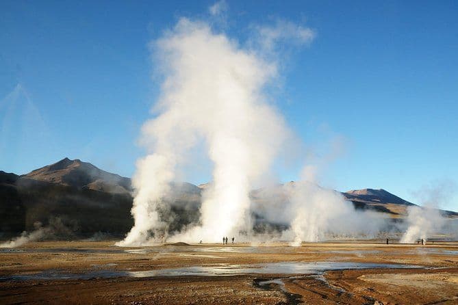 Un voyage en groupe WeRoad observe de grands geysers crachant de la vapeur dans une vaste vallée aride avec des montagnes en arrière-plan.