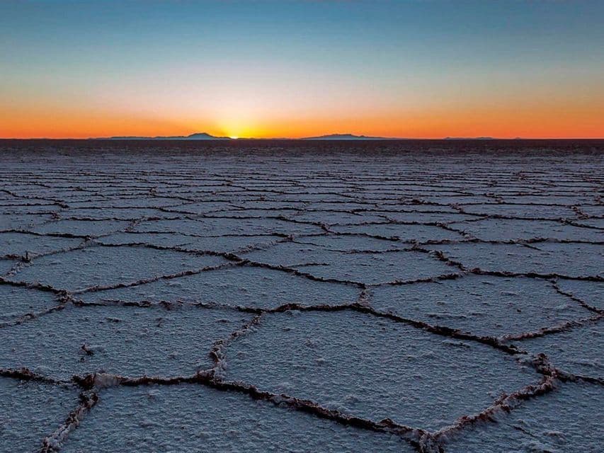 Il sole tramonta all'orizzonte su una vasta salina con un motivo geometrico e screpolato sul terreno.