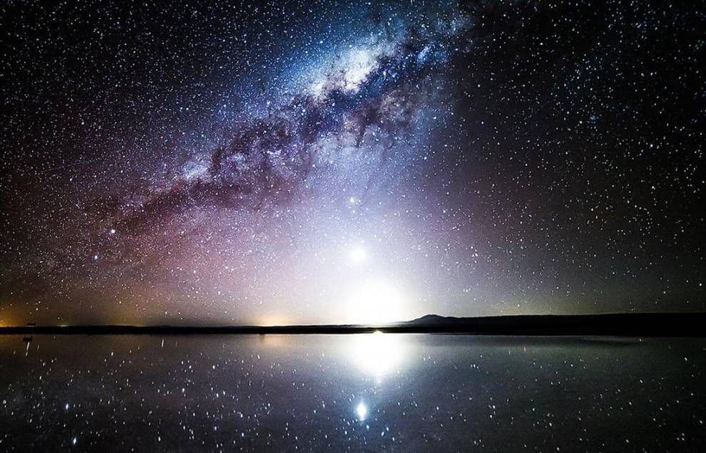 The Milky Way and countless stars in the night sky perfectly reflect onto a calm body of water on the horizon.