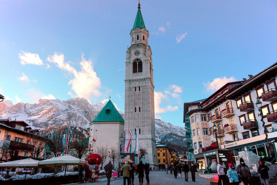 Un alto campanile in pietra con una guglia verde si staglia in una piazza di paese animata da persone, con lo sfondo di montagne innevate.
