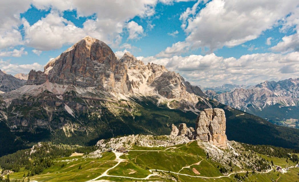 Una vista panoramica di una catena montuosa rocciosa, con le Cinque Torri che si stagliano su verdi colline e sentieri sinuosi.