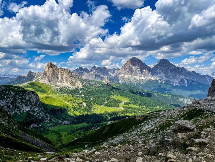 Una vista panoramica di una catena montuosa rocciosa che domina una valle verde lussureggiante, il tutto sotto un cielo dinamico con nuvole bianche e soffici.