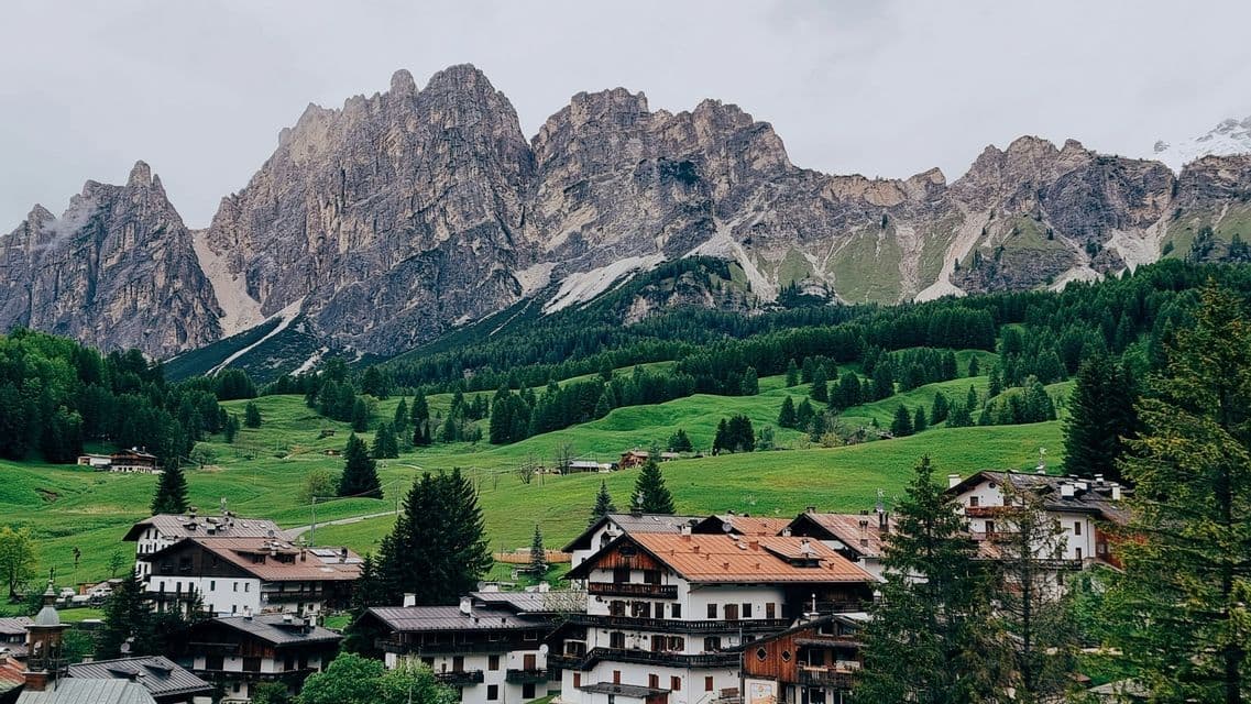 Un villaggio alpino con case tradizionali si adagia in una lussureggiante valle verde ai piedi di una catena montuosa frastagliata sotto un cielo nuvoloso.