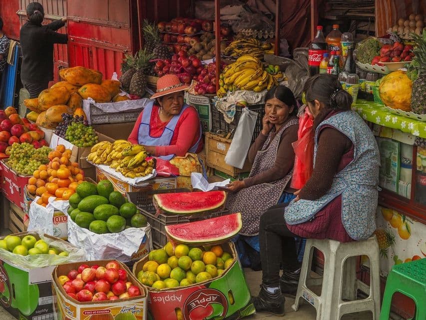 Trois vendeuses discutent assises devant un étal de marché extérieur coloré, regorgeant de fruits frais variés.