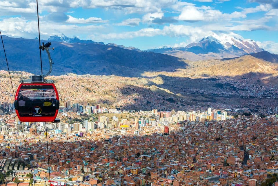 A red cable car glides over a sprawling city nestled in a valley, with large snow-capped mountains in the background.