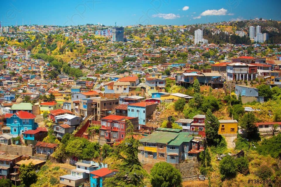 Eine dichte Stadtlandschaft mit farbenfrohen Häusern und Gebäuden erstreckt sich über einen weitläufigen grünen Berghang unter einem klaren blauen Himmel.