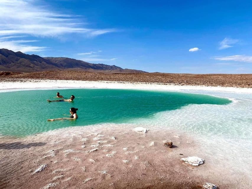 A WeRoad group trip of three people floating in a turquoise salt lake, surrounded by a desert landscape and mountains.