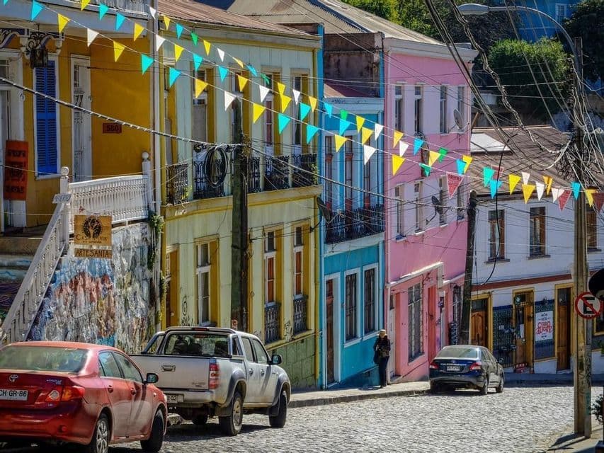 Des guirlandes de drapeaux colorés flottent au-dessus d'une rue pavée bordée de bâtiments multicolores vifs et de voitures garées.