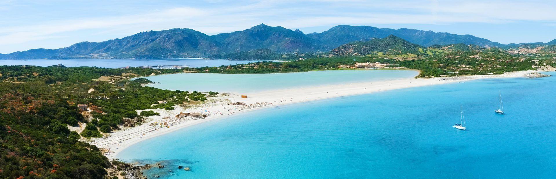 Una vista aérea panorámica de una larga playa de arena blanca que separa aguas turquesas, con dos veleros y una costa montañosa al fondo.