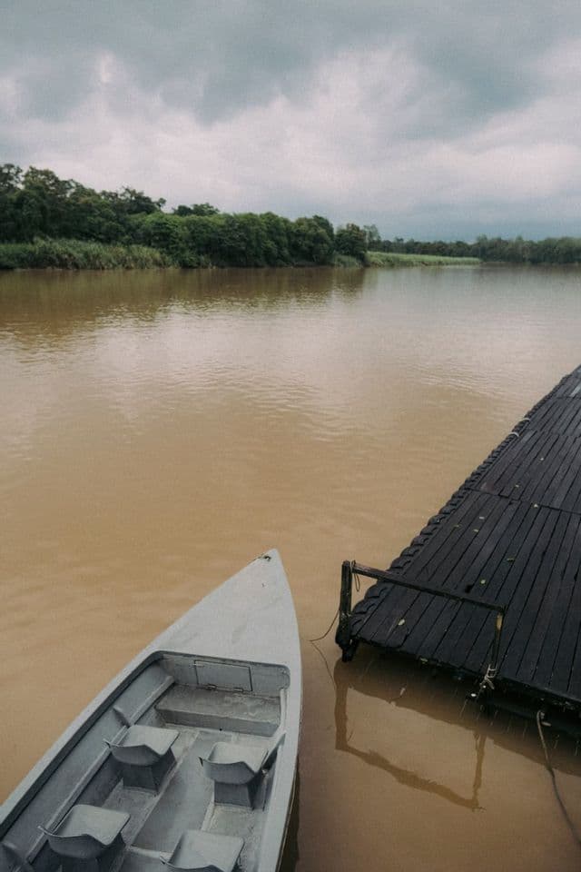 Un pequeño bote flota junto a un oscuro muelle de madera en un río ancho y turbio bajo un cielo nublado.