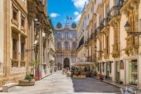 Una strada stretta e soleggiata, fiancheggiata da edifici storici, conduce a un grande arco con una bandiera italiana in cima.
