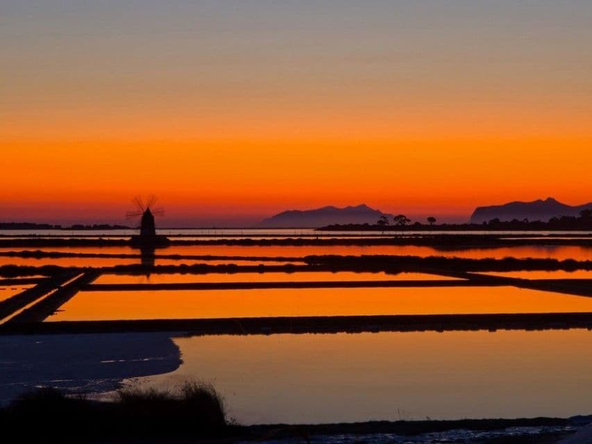 Una sagoma di mulino a vento si staglia tra le saline, riflettendo un vivace tramonto arancione, con montagne lontane all'orizzonte.