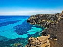 Scogliere rocciose si affacciano su uno specchio d'acqua con vivaci sfumature di turchese e blu intenso sotto un cielo sereno.