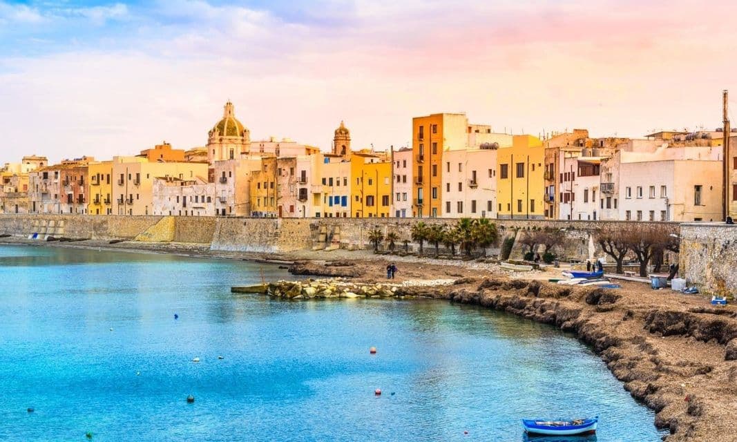 Una cittadina costiera con edifici colorati si affaccia su una diga di pietra lungo acque di un blu brillante, sotto un cielo dalle sfumature pastello.
