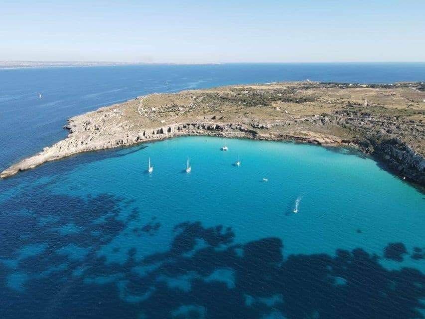 Vista aerea di barche a vela che galleggiano in una baia turchese e cristallina, accanto a una penisola rocciosa e arida sotto un cielo blu.