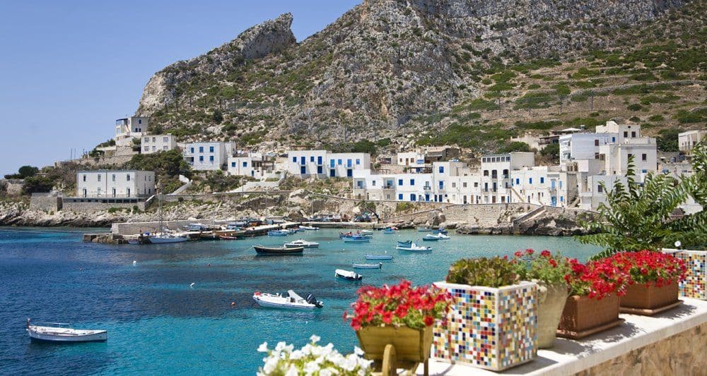 Un balcone con fiori in vaso si affaccia su un villaggio costiero con edifici bianchi e barche in una baia turchese sotto un cielo sereno.