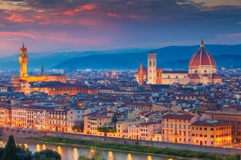 Uno skyline cittadino storico con una grande cupola e un'alta torre, illuminato al tramonto e un fiume in primo piano.