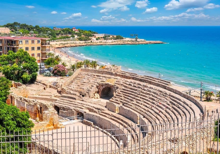 Un antico anfiteatro in pietra si affaccia su una spiaggia sabbiosa e un mare turchese, con una città costiera sullo sfondo sotto un cielo blu.