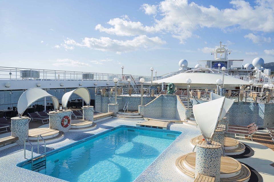 Piscina vuota sul ponte piastrellato di una nave da crociera, con aree relax e tendalini sotto un cielo azzurro.