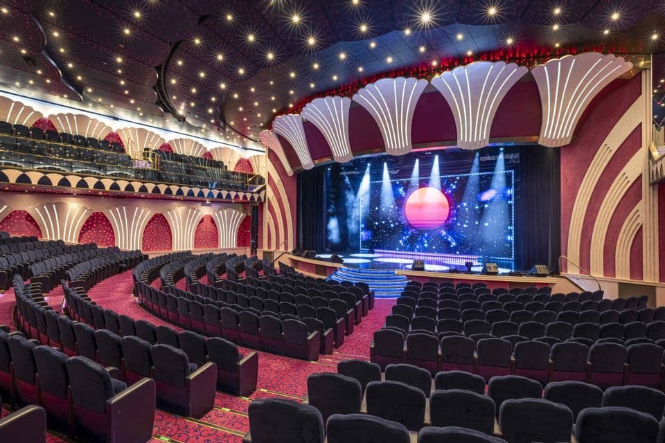 Un'ampia inquadratura di un teatro vuoto e ornato, con file curve di poltrone e un palcoscenico illuminato a giorno.
