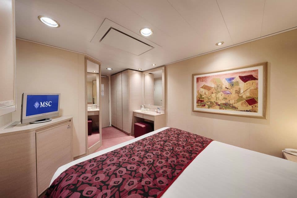 Interno di una cabina di nave da crociera MSC, con letto, runner a fantasia rossa, toeletta, TV e armadi.