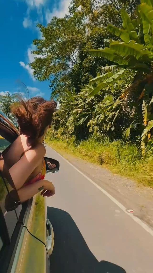 Una donna con i capelli che sventolano nel vento si sporge dal finestrino di un'auto in corsa, accanto a una strada costeggiata da vegetazione tropicale.
