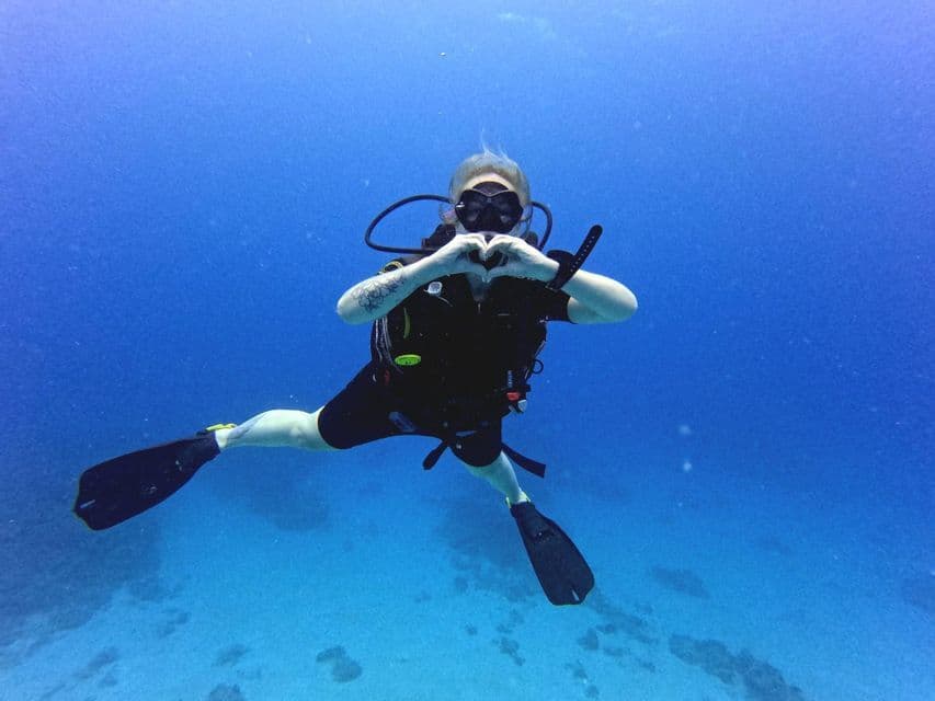 Un plongeur flotte dans des eaux bleues cristallines, formant un cœur avec ses mains vers l'appareil photo.