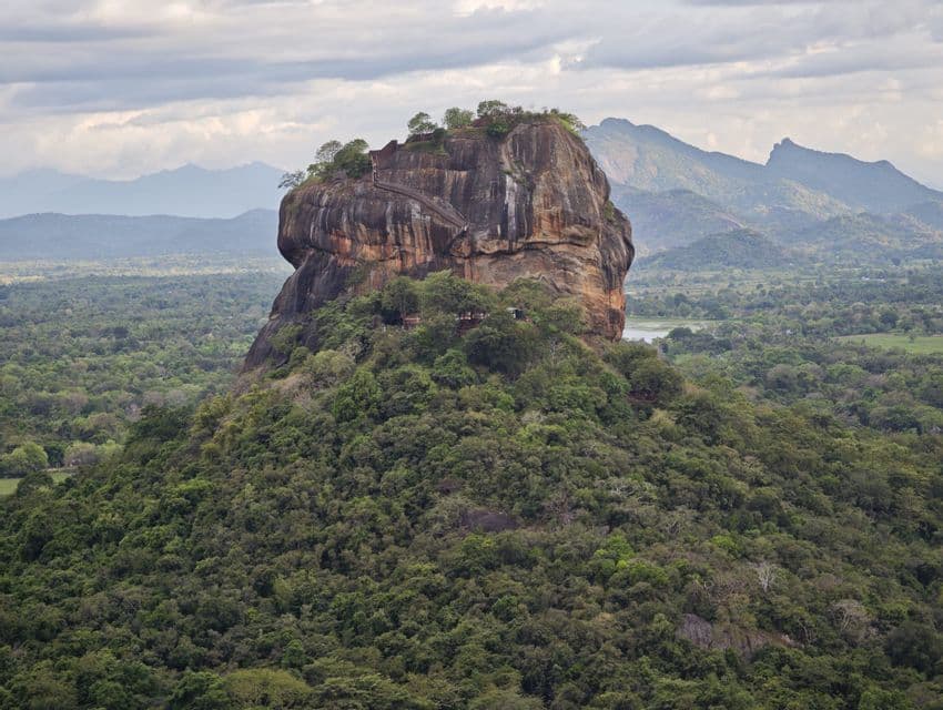 Une grande forteresse rocheuse s'élève d'une jungle verte dense, avec une chaîne de montagnes visible à l'arrière-plan sous un ciel nuageux.