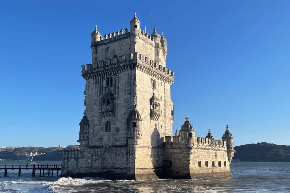 An ornate, fortified stone tower stands in the water under a clear blue sky, with gentle waves lapping at its base.