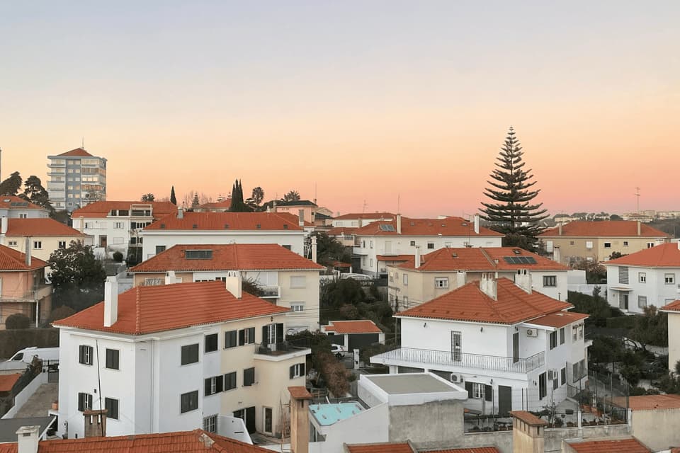 A cluster of white houses with red-tiled roofs on a hillside under a warm, pastel-colored sunset sky.