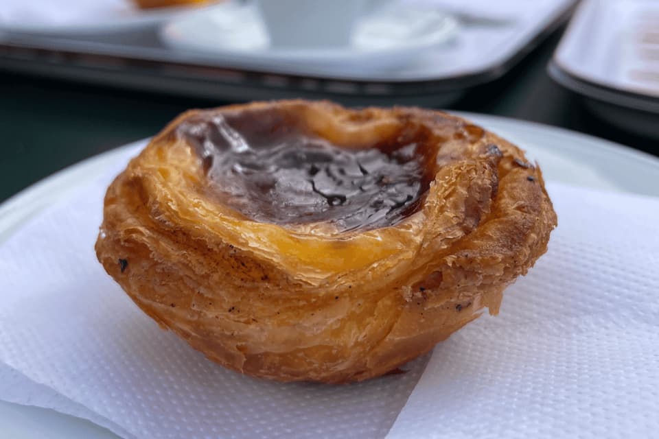 A close-up of a pastel de nata, a Portuguese egg tart, with a flaky crust and caramelized top, sitting on a white napkin.