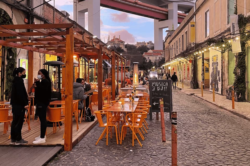People dining at an outdoor restaurant with orange chairs on a cobblestone street under a large bridge at dusk.