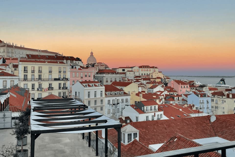A cityscape of colorful buildings with red-tiled roofs viewed from a terrace at sunset over the water.