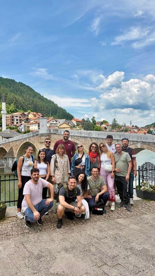 Un viaggio di gruppo WeRoad posa per una foto di fronte a un grande ponte di pietra, con una città e una collina verde sullo sfondo.