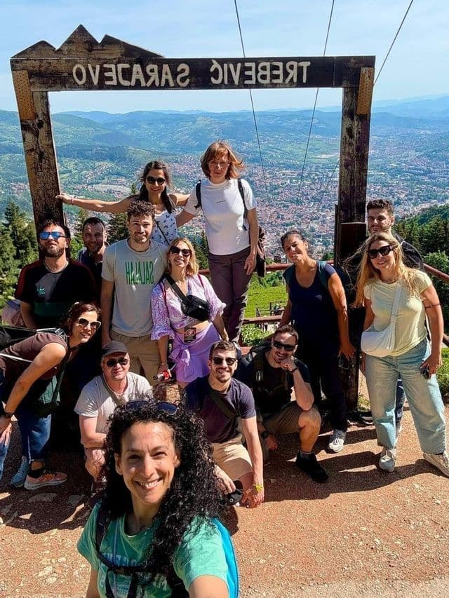 Un viaggio di gruppo WeRoad scatta un selfie su un punto panoramico in montagna, con una città e colline visibili sullo sfondo.
