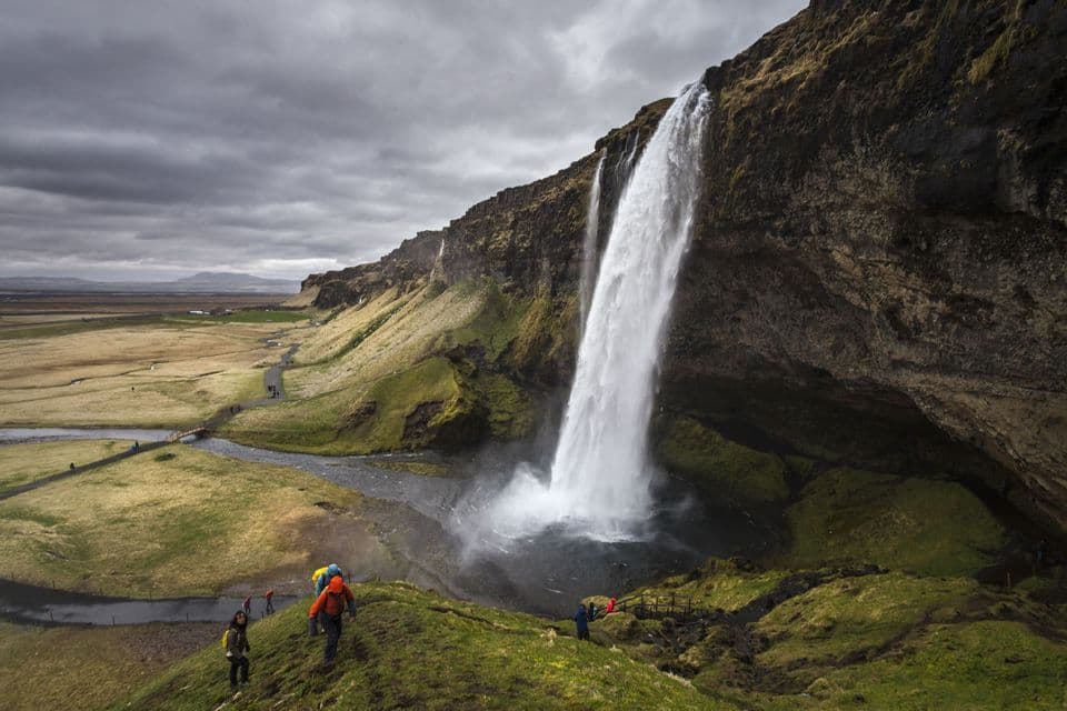 A WeRoad group trip hikes on a green hill overlooking a large waterfall flowing down a rocky cliff into a wide valley.