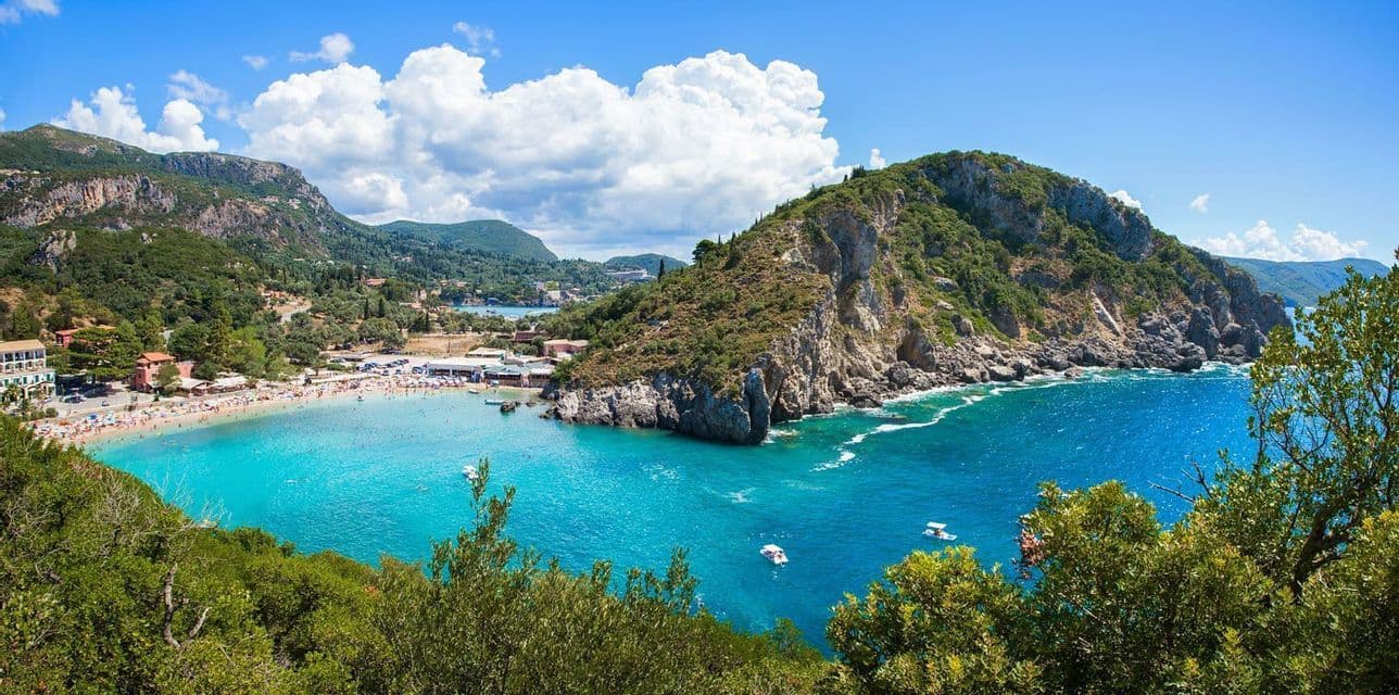 An elevated view of a busy beach with turquoise water in a cove, surrounded by tree-covered cliffs and mountains.