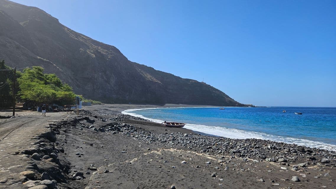 Ein weiter Blick auf einen schwarzen Sandstrand mit einem Boot am Ufer, türkisfarbenem Wasser und einem großen Hügel unter klarem Himmel.