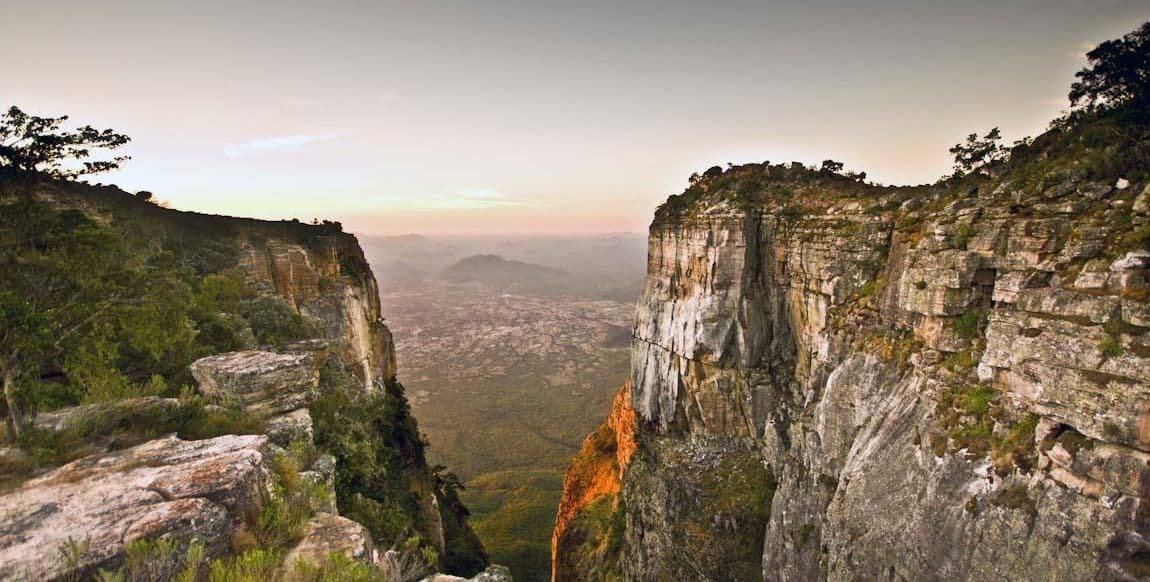 Una vista tra due scogliere a picco che si affacciano su una vasta valle e una città lontana all'alba.