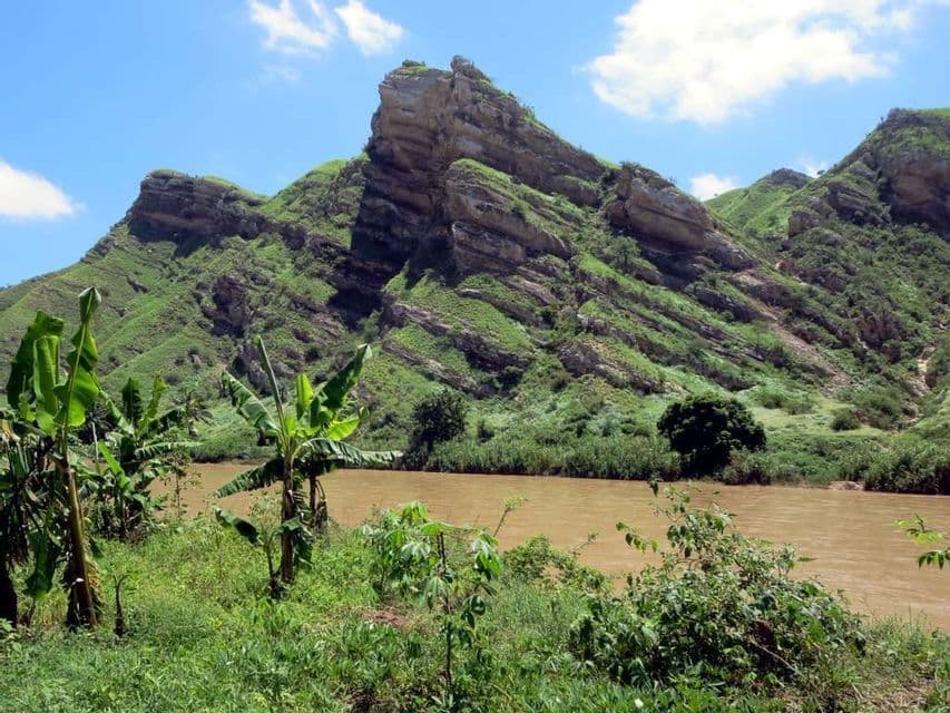 Un fiume fangoso scorre lungo una rigogliosa sponda verde con banani, con grandi montagne verdi stratificate sullo sfondo sotto un cielo azzurro.