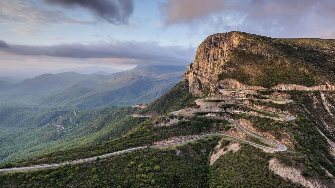 Veduta aerea di una strada sinuosa con molti tornanti che si inerpica su una ripida montagna, coperta di vegetazione verde, sotto un cielo nuvoloso.