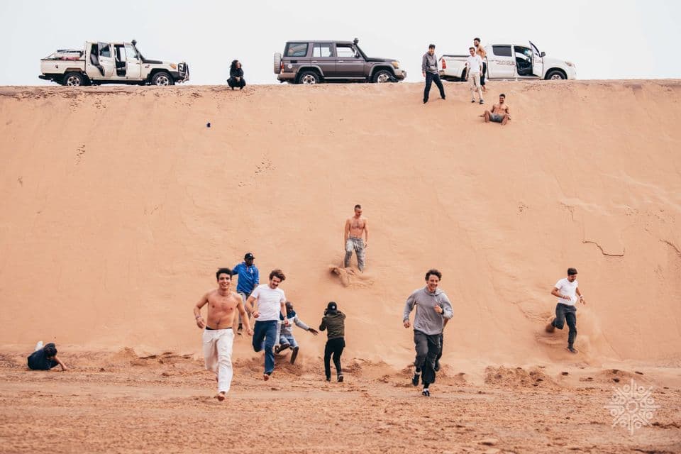 Un viaggio di gruppo WeRoad con persone che corrono e scivolano giù da una grande duna di sabbia, con veicoli fuoristrada parcheggiati in cima.