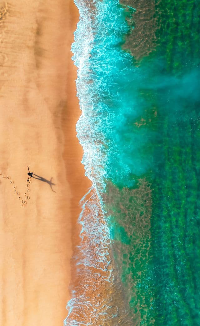 Una vista aerea dall'alto di una persona che cammina su una spiaggia sabbiosa accanto all'acqua turchese, con impronte che si lasciano alle spalle.