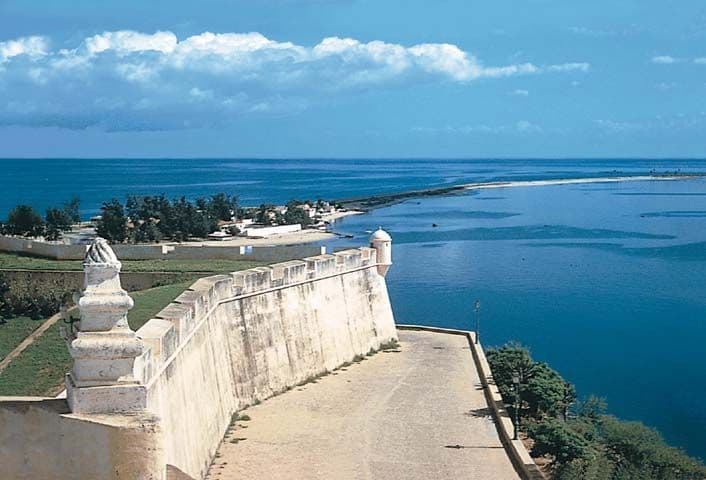 Una vista da una muraglia di fortezza in pietra con una torre di guardia, affacciata su un mare blu calmo e una barra di sabbia sotto un cielo parzialmente nuvoloso.