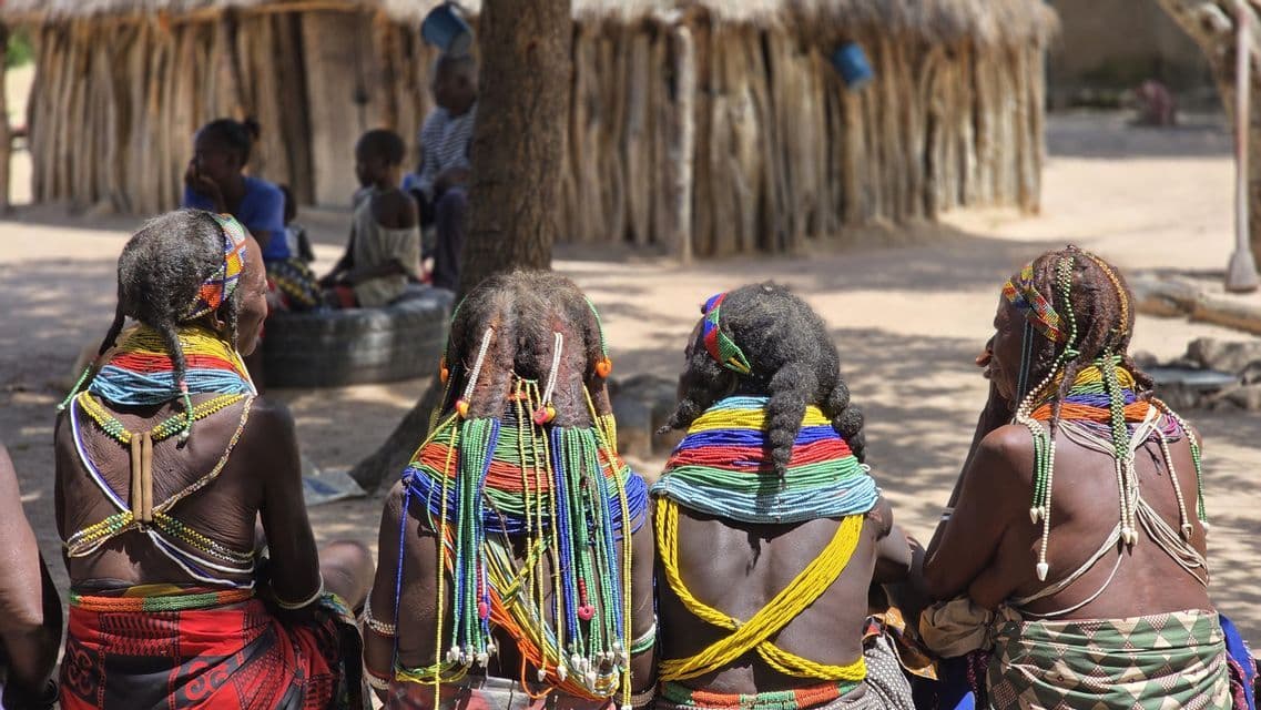 Quattro donne con capelli adorni di perline e collane sono sedute insieme, viste da dietro in un villaggio.