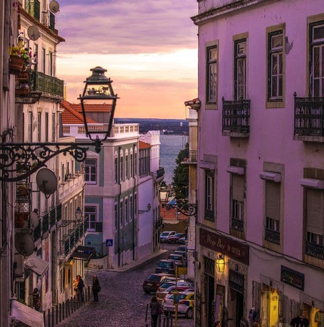 A cobblestone street slopes downhill between colorful buildings, with a view of the water at sunset.