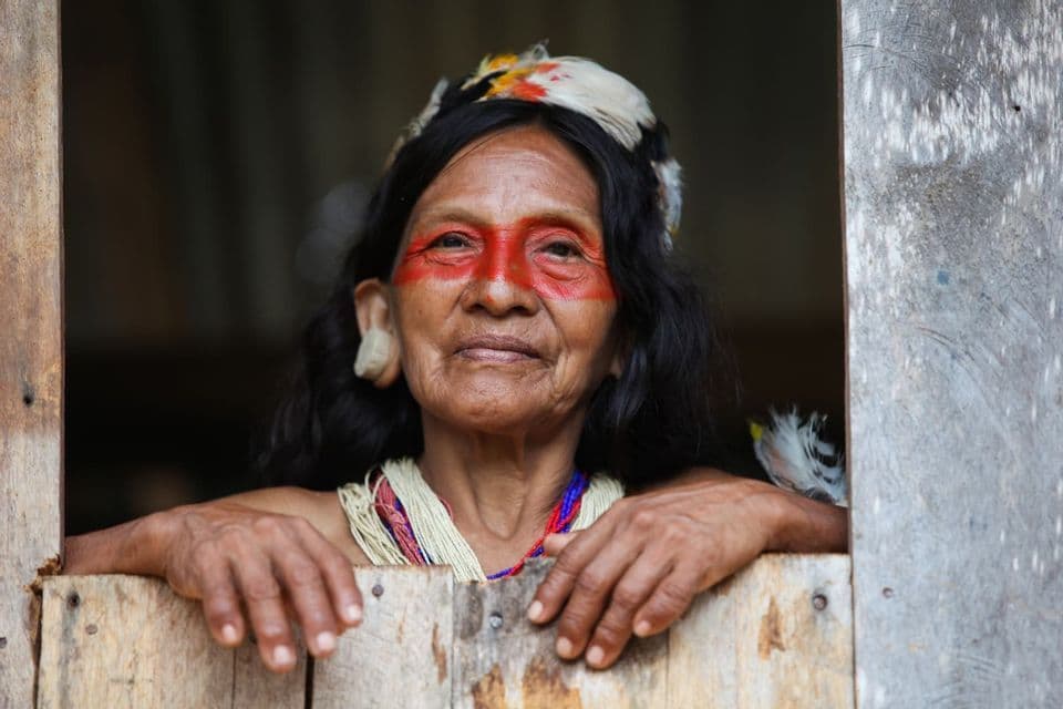 Un'anziana donna indigena con il viso dipinto di rosso e un copricapo di piume guarda attraverso una cornice di legno.