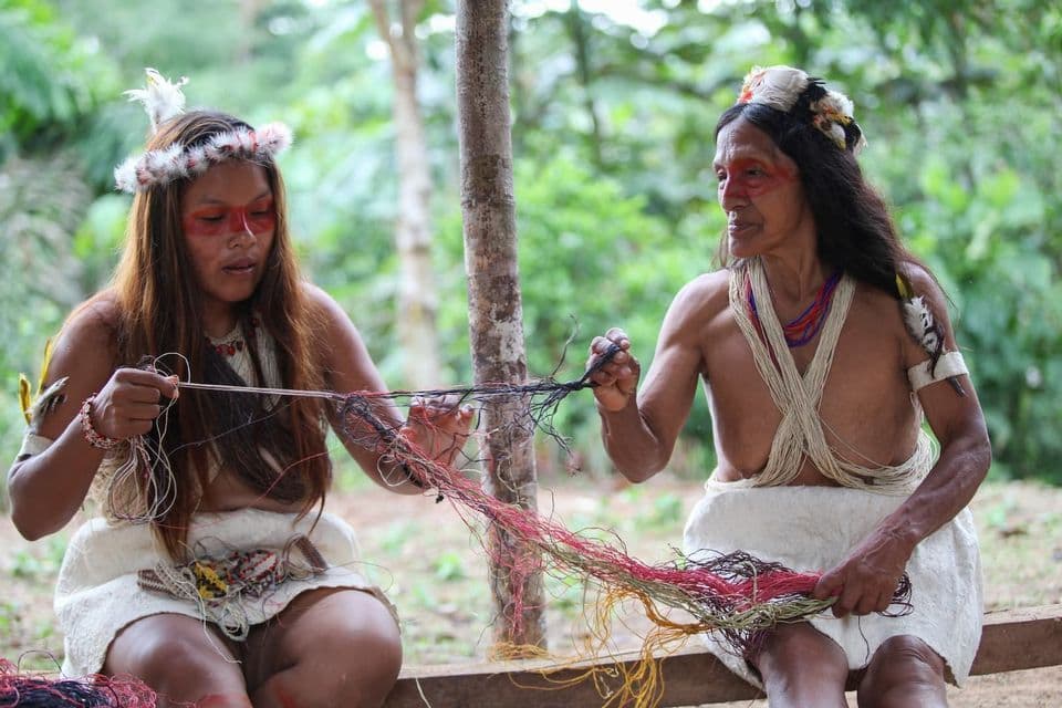 Two indigenous women in traditional clothing and feather headdresses work together with colorful threads outdoors.